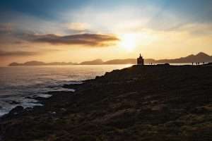 Imagen de un atardecer en una playa de Galicia, con la silueta de un faro en el horizonte.
